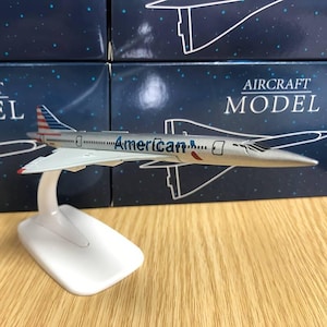 Può includere: Un modello di aereo Concorde argentato e blu su un supporto bianco. L'aereo ha la scritta "American..." sul lato. L'aereo è su una superficie di legno con scatole sullo sfondo.