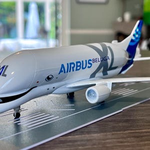 May include: A white model Airbus Beluga XL aircraft with blue tail detailing and the Airbus and Beluga XL logos. The model is positioned on a runway-style surface, showcasing the plane's design.