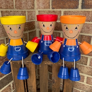 May include: Three painted terracotta pots shaped like scarecrows with blue overalls, yellow, red, and orange hats, and blue bell bottoms. The scarecrows are sitting on a wooden crate.