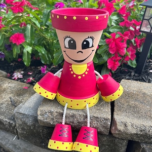 May include: A pink and yellow painted terracotta planter shaped like a person with a smiling face, wearing a red dress, yellow necklace, and red shoes with yellow polka dots.