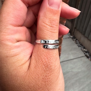 May include: A silver ring with the engraved words "olite fe bastardes carborundum". The ring is wrapped around a finger. The ring has a simple, elegant design. The ring is made of a shiny metal.