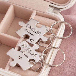 May include: Two silver puzzle piece keyrings with engraved text and heart charms. Each keyring has a date and name: "23.06.20 Laura" and "23.06.20 James". The keyrings are displayed in a pink jewellery box.