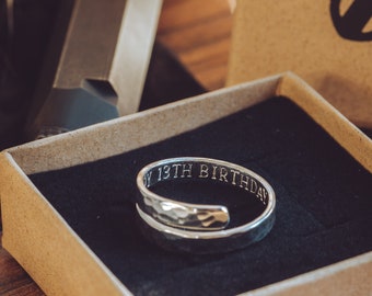 May include: A silver ring with a hammered finish and the inscription "My 13th Birthday" on the band. The ring is displayed in a brown box with a black velvet lining.