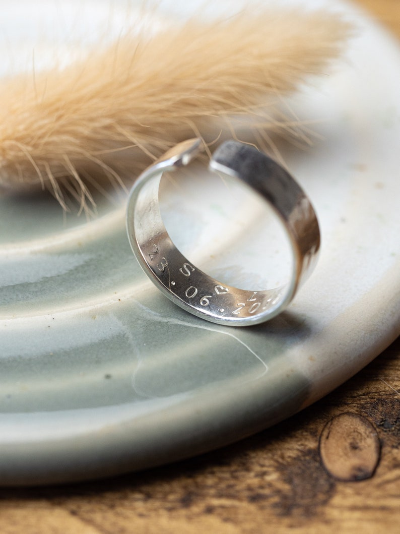 May include: A silver ring with a hammered finish and a personalized inscription that reads "28.06.2021". The ring is resting on a light blue ceramic dish.