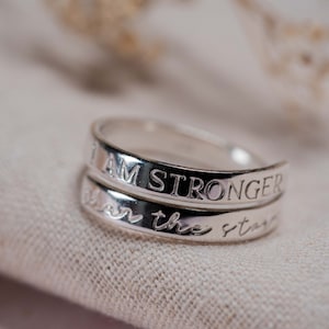 May include: Two silver rings stacked on top of each other. The top ring has the words "I AM STRONGER" engraved on it. The bottom ring has the words "than the stars" engraved on it.