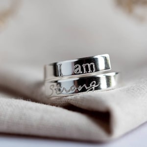 May include: Close-up of two silver rings, each engraved with the words "I am Strong". The rings are intertwined and sit on a beige cloth. The rings have a shiny, reflective surface.