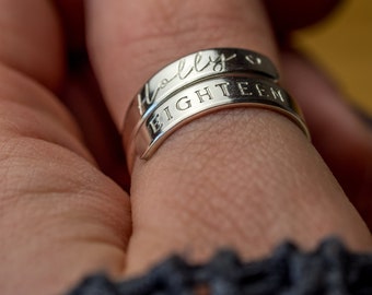 May include: Two silver rings on a finger. The top ring has the name "Holly" engraved on it. The bottom ring has the word "EIGHTEEN" engraved on it.