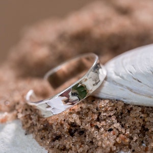 May include: A silver hammered ring with a smooth, rounded surface. The ring is sitting on a bed of sand and a white seashell.
