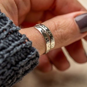 May include: Two silver rings with a star pattern on the band. The rings are worn on the thumb of a hand.