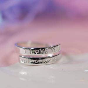 May include: Silver ring with the words "I love you" and "Mommy" engraved on it. The ring is wrapped around itself and has a small heart symbol on each inscription.