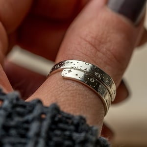 May include: A silver ring with a textured design of stars and circles. The ring is worn on a person's thumb.