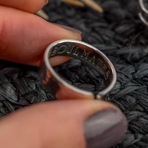 May include: A silver ring with the inscription "SOULMATES" engraved on the inside. The ring is held between fingers, displaying the text. The ring has a simple band design, reflecting a minimalist aesthetic.