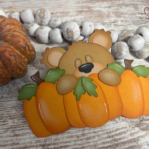 May include: A die-cut paper craft featuring a brown bear peeking over three orange pumpkins with green leaves. The bear has a black nose and closed eyes. The design is layered with textured cardstock, showcasing a whimsical autumn theme with a warm color palette of orange, brown, and green tones, suitable for seasonal scrapbooking or card making projects.