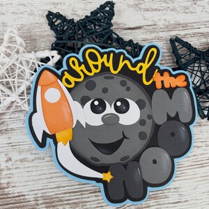 May include: A whimsical, layered papercraft featuring a smiling gray moon with a rocket ship and the words "around the moon" in yellow and orange. The design is outlined in blue and includes a small yellow star. Decorative star accents are in the background.