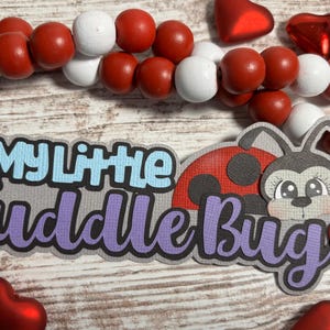 May include: A decorative sign with the text "My Little Cuddle Bug" in blue and purple, featuring a ladybug and bunny design. Red and white beads and heart-shaped decorations surround the sign.