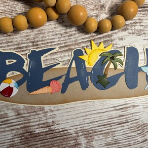 May include: A decorative sign spelling "BEACH" in blue letters with beach-themed embellishments. Includes a beach ball, seashell, sun, palm tree, and umbrella. The sign is on a light brown background, with a wooden bead garland above.