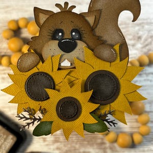 May include: A brown squirrel-shaped papercraft figure holding three yellow sunflowers with brown centers. The squirrel has large eyes and a black nose. The sunflowers have green leaves and are arranged in a bouquet.