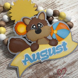 May include: A brown squirrel with big eyes holds a beach ball and a banner that says "August" in blue letters. The squirrel is surrounded by yellow and brown beads.