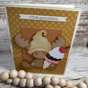May include: A handmade greeting card with a brown and tan moose wearing an ice cream cone hat. The card has the text "CONE-gratulations" and a decorative ice cream cone with a cherry on top.