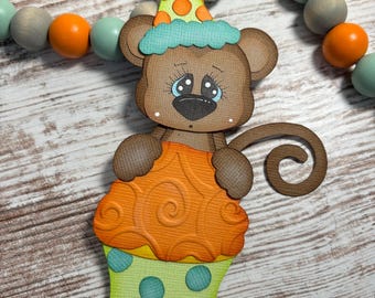 May include: A brown bear-shaped paper cutout wearing a party hat and holding an orange cupcake. The cupcake has a yellow base with blue polka dots. A string of wooden beads in orange, gray, and teal is in the background.