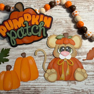 May include: A set of fall-themed paper crafts featuring a pumpkin patch sign, a mouse wearing a pumpkin hat, and two pumpkins. The mouse is holding a pumpkin in its paws and has a yellow ribbon around its neck. The pumpkins are orange and have green stems.