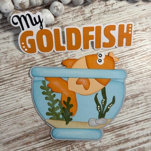 May include: A layered papercraft design featuring a goldfish in a blue fishbowl. The orange goldfish is partially visible, with the text "My GOLDFISH" above. The design includes green seaweed and small pebbles.