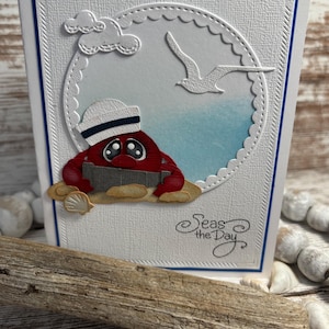 May include: A handmade greeting card with a nautical theme. It features a red crab wearing a sailor hat, a seagull, and the phrase "Seas the Day." The background has a blue watercolor effect, and the card is framed with a blue border.
