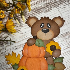 May include: A brown bear cutout holding an orange pumpkin and a yellow sunflower. The bear has large eyes and a black nose. The image is decorated with fall leaves and flowers, set against a wood-grain background.
