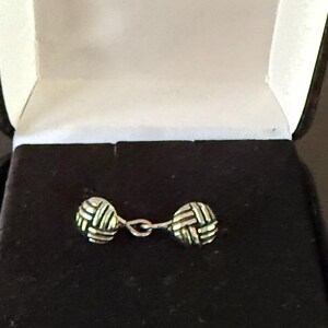 May include: A pair of silver cufflinks with a woven design, presented in a black velvet box with a white interior. The cufflinks are connected by a delicate chain, and the box is open.