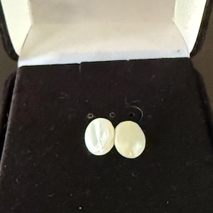 May include: A pair of oval-shaped, off-white earrings displayed inside a black velvet jewelry box with a white interior. The earrings are positioned in the center of the box, showcasing their simple elegance. The box is open, revealing the earrings.