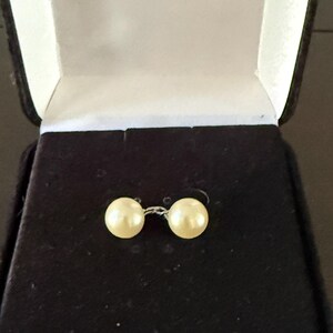 May include: A pair of cream-colored pearl earrings displayed in an open black velvet jewelry box with a white satin interior. The earrings are connected by a thin silver wire.