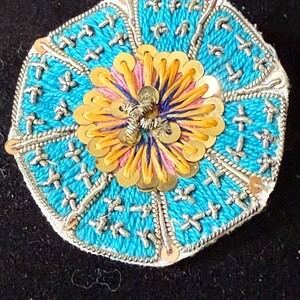 May include: A round, embroidered embellishment with a central flower design. The flower features yellow sequins and pink and blue thread. The outer ring is turquoise with silver cross-stitch details and a silver border. The background is black.