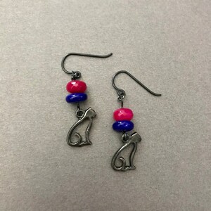 May include: A pair of dangle earrings featuring a cat silhouette charm. Each earring has a dark metal hook, with a stack of a pink and blue bead. The earrings are displayed on a light gray surface.