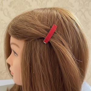 May include: A rectangular hair clip with a textured, bright red surface. The clip is secured horizontally into light brown hair, holding a small section of hair back from the side of the head. The accessory features a simple, flat design.