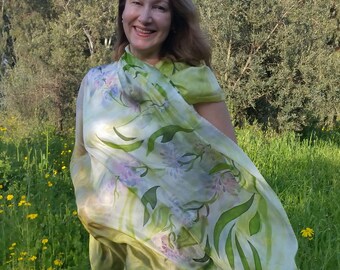 May include: A woman wearing a flowing, sheer scarf with a floral design in shades of green, white, and purple. The scarf is draped over the shoulders and arms. The background is a field of green grass and yellow flowers.