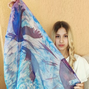 May include: A blue and purple silk scarf with a white background and a design of flying birds. The scarf is being held by a person with long blonde hair.