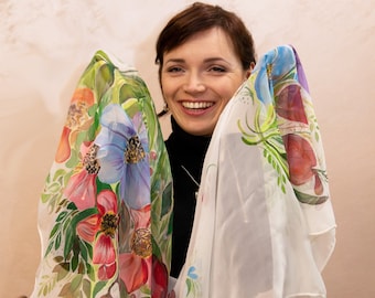 Könnte beinhalten: Eine Frau in einem schwarzen Rollkragenpullover hält zwei weiße Seidentücher mit Blumendesigns. Die Tücher sind mit bunten Blumen und Blättern bemalt.