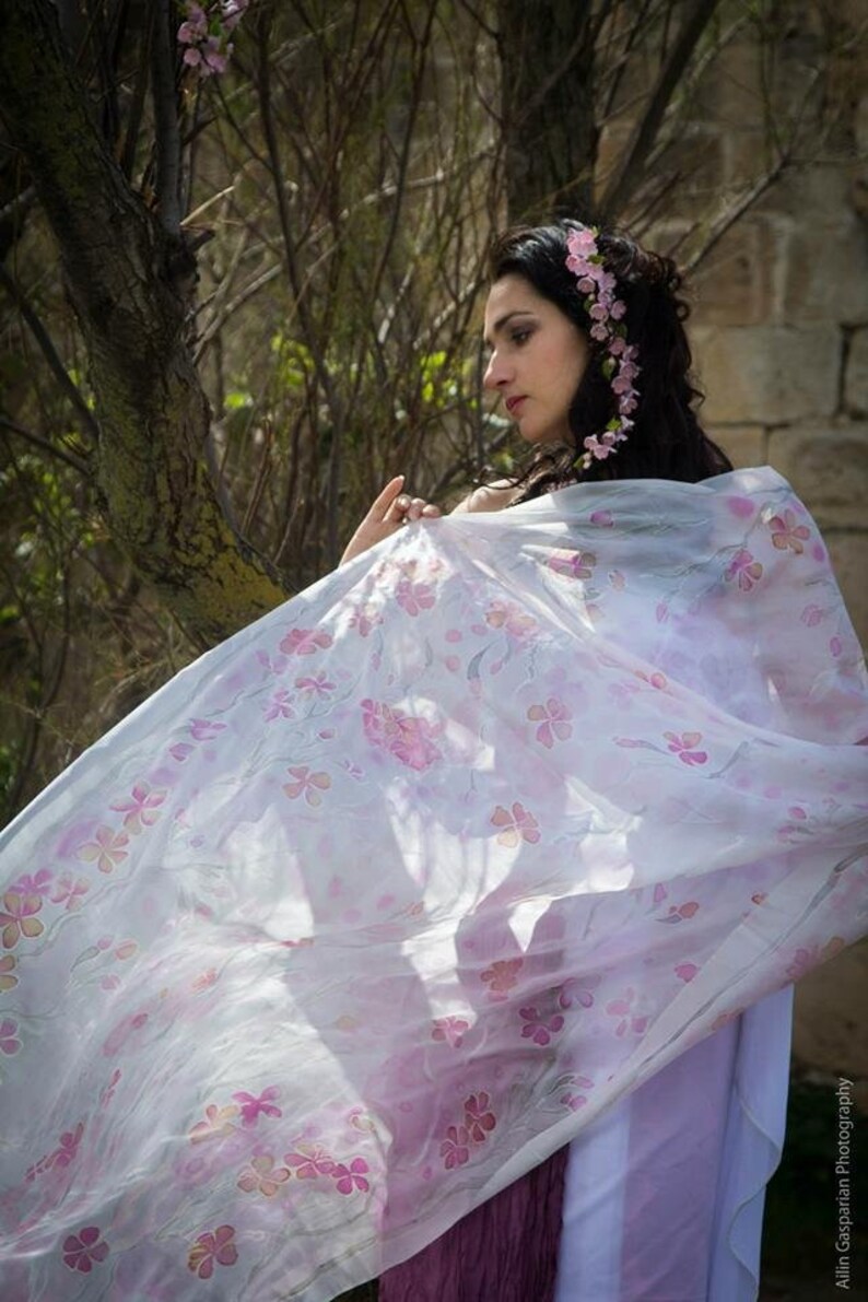 May include: A woman wearing a white sheer scarf with pink floral designs. The scarf is flowing in the wind. She is standing in front of a tree with pink blossoms.