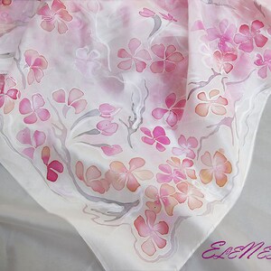 May include: A white silk scarf with a delicate pink floral pattern. The flowers are painted in shades of pink and orange, and the branches are a light gray. The scarf is a beautiful and elegant accessory.
