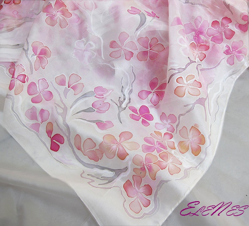 May include: A white silk scarf with a delicate pink floral pattern. The flowers are painted in shades of pink and orange, and the branches are a light gray. The scarf is a beautiful and elegant accessory.