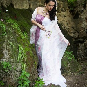 May include: A woman in a white and purple dress with floral accents stands in a mossy, rocky area. She holds a small bouquet of pink flowers in her hand.