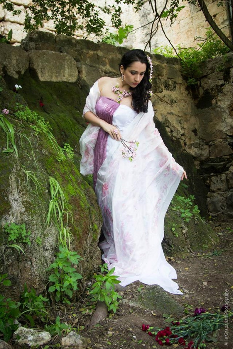 May include: A woman in a white and purple dress with floral accents stands in a mossy, rocky area. She holds a small bouquet of pink flowers in her hand.