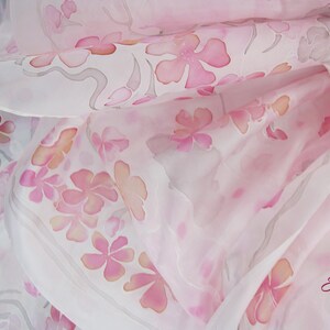 May include: A white silk scarf with a delicate pink floral pattern. The scarf is made of soft, flowing silk and features a subtle ombre effect.