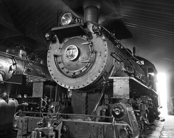 May include: A black and white photo of a vintage steam locomotive with the number 475 on the side. The locomotive is in a train shed with a bright light shining through the open doors.