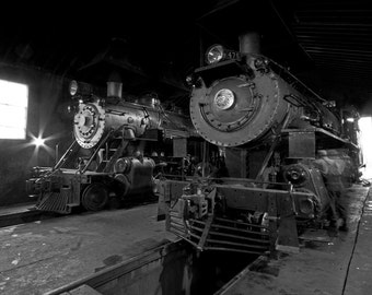 May include: Black and white image of two vintage steam locomotives inside a dimly lit train shed. The locomotives are large, with prominent wheels, boilers, and smokestacks. One locomotive has the number 475 displayed on its front.