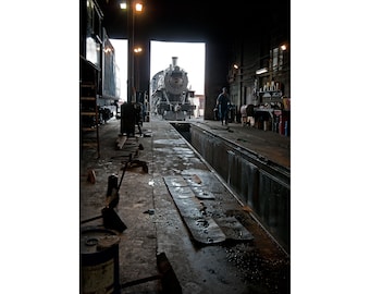 May include: A black steam locomotive is being worked on in a large, dark, industrial building. The locomotive is partially inside the building and partially outside. The building is lit by overhead lights.