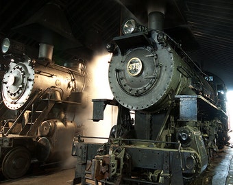 May include: Two vintage steam locomotives, one labeled "475" and the other "90", are parked in a dimly lit train shed. Both locomotives are black and have large wheels. Steam is rising from the locomotives, creating a hazy atmosphere.