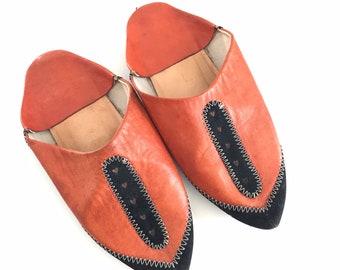 May include: A pair of brown leather slippers with black trim and a pointed toe. The slippers have a decorative heart pattern on the trim.