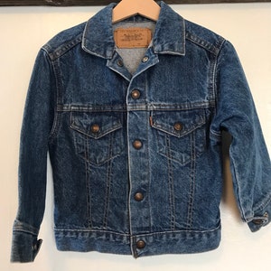 K&ouml;nnte beinhalten: Eine blaue Jeansjacke mit einem braunen Lederetikett, auf dem "Levi's Strauss & Co." steht. Die Jacke hat einen Knopfverschluss und zwei Fronttaschen.