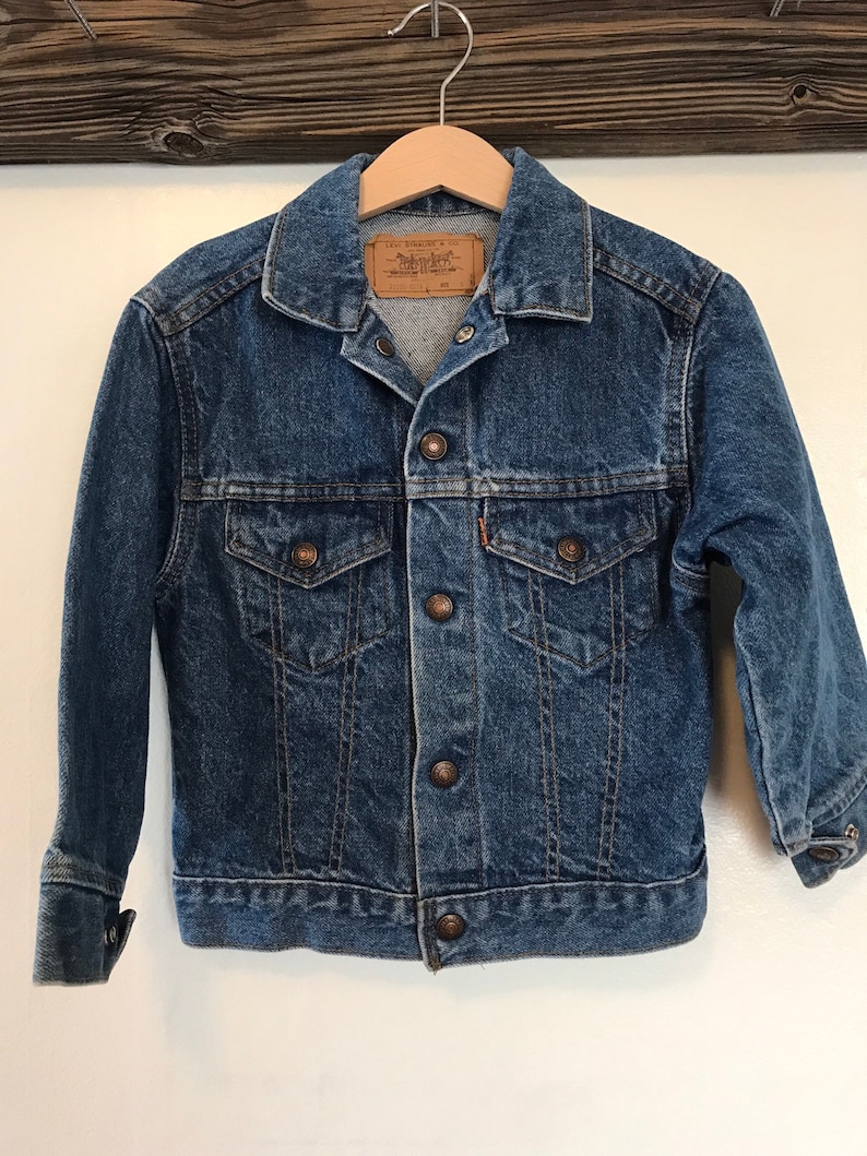 K&ouml;nnte beinhalten: Eine blaue Jeansjacke mit einem braunen Lederetikett, auf dem "Levi's Strauss & Co." steht. Die Jacke hat einen Knopfverschluss und zwei Fronttaschen.